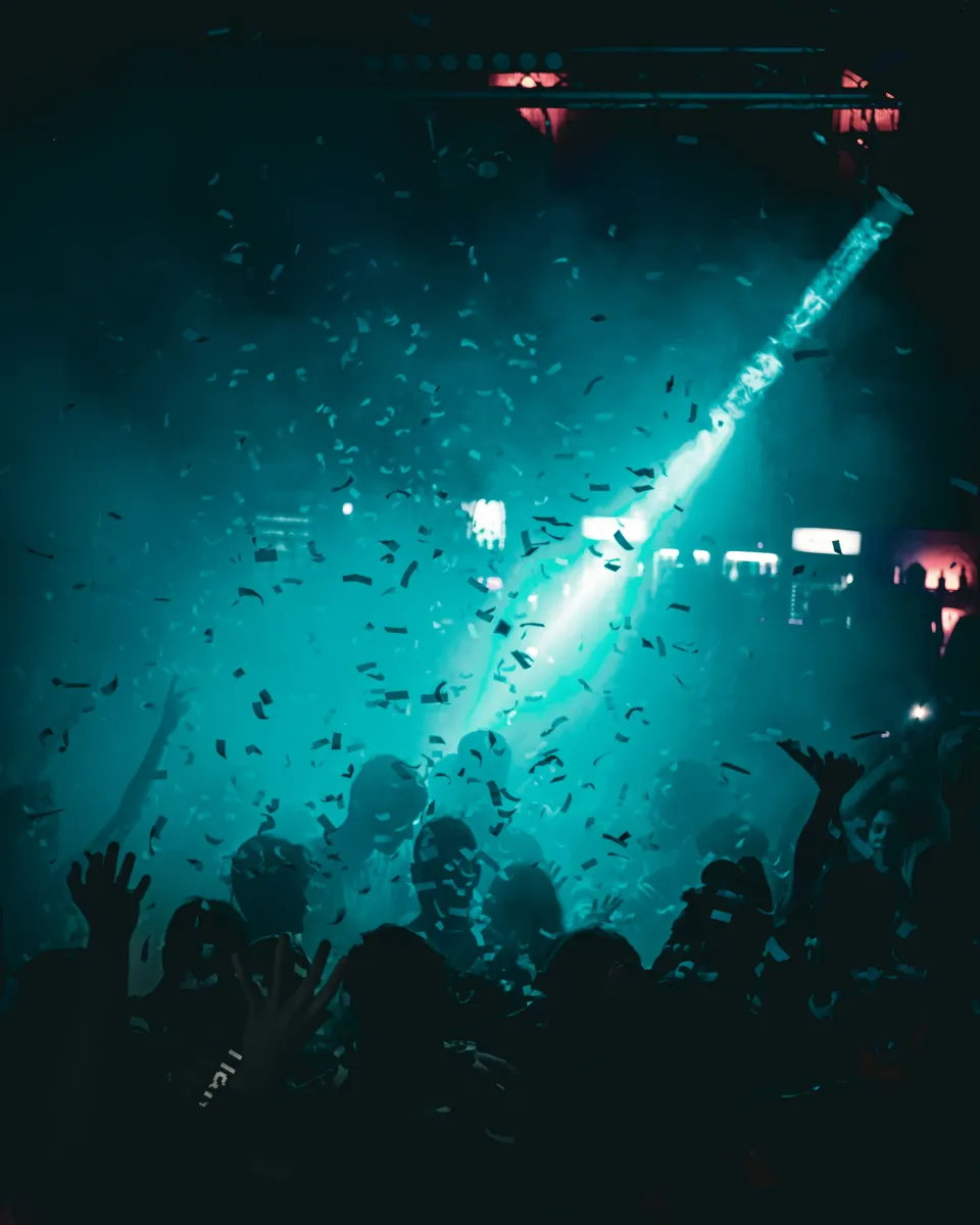 Une foule mains levées sous une pluie de confettis, traversée par un faisceau de lumière.