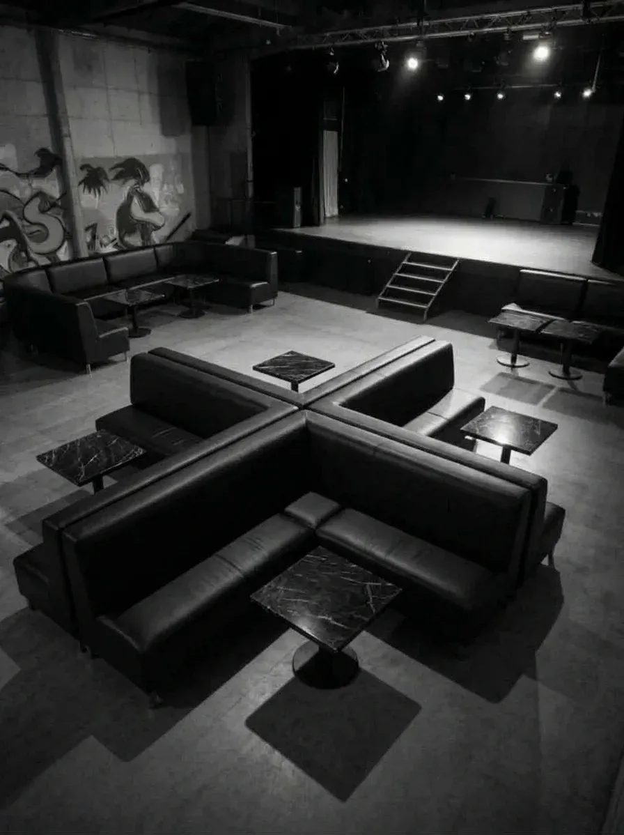 La salle vide en noir et blanc, banquettes de cuir en croix autour de tables de marbre, la scène au fond.