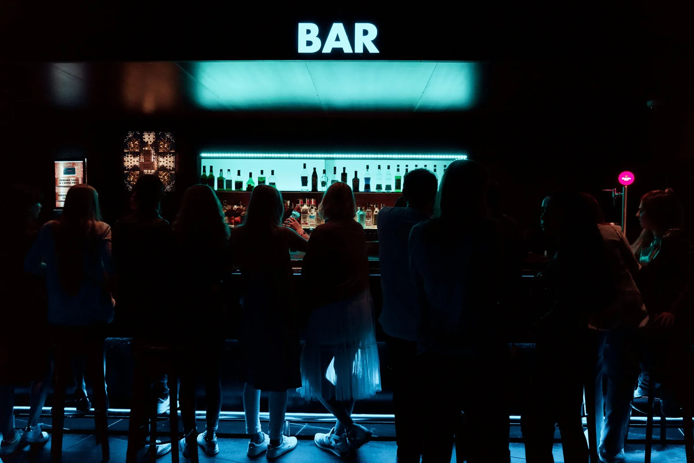 Le comptoir du bar sous son enseigne lumineuse, des personnes accoudées de dos.
